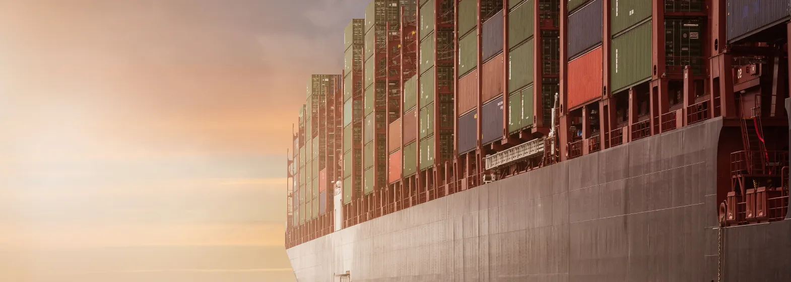 Close-up of the side of a large cargo ship stacked with multicolored shipping containers against a hazy sky at sunset.